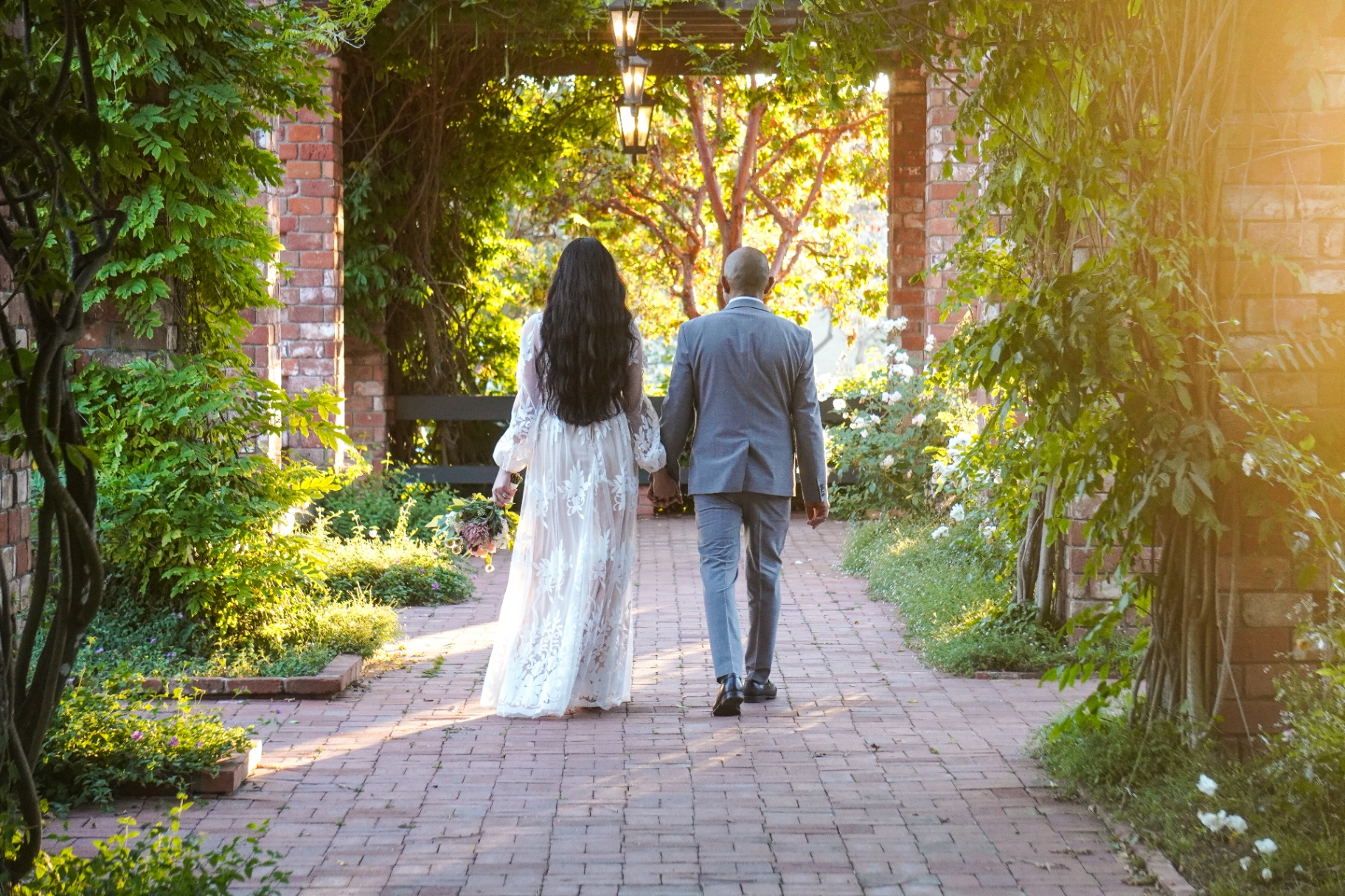 Bride and groom walking hand in hand through a sunlit rose-garden pergola at a Santa Barbara wedding