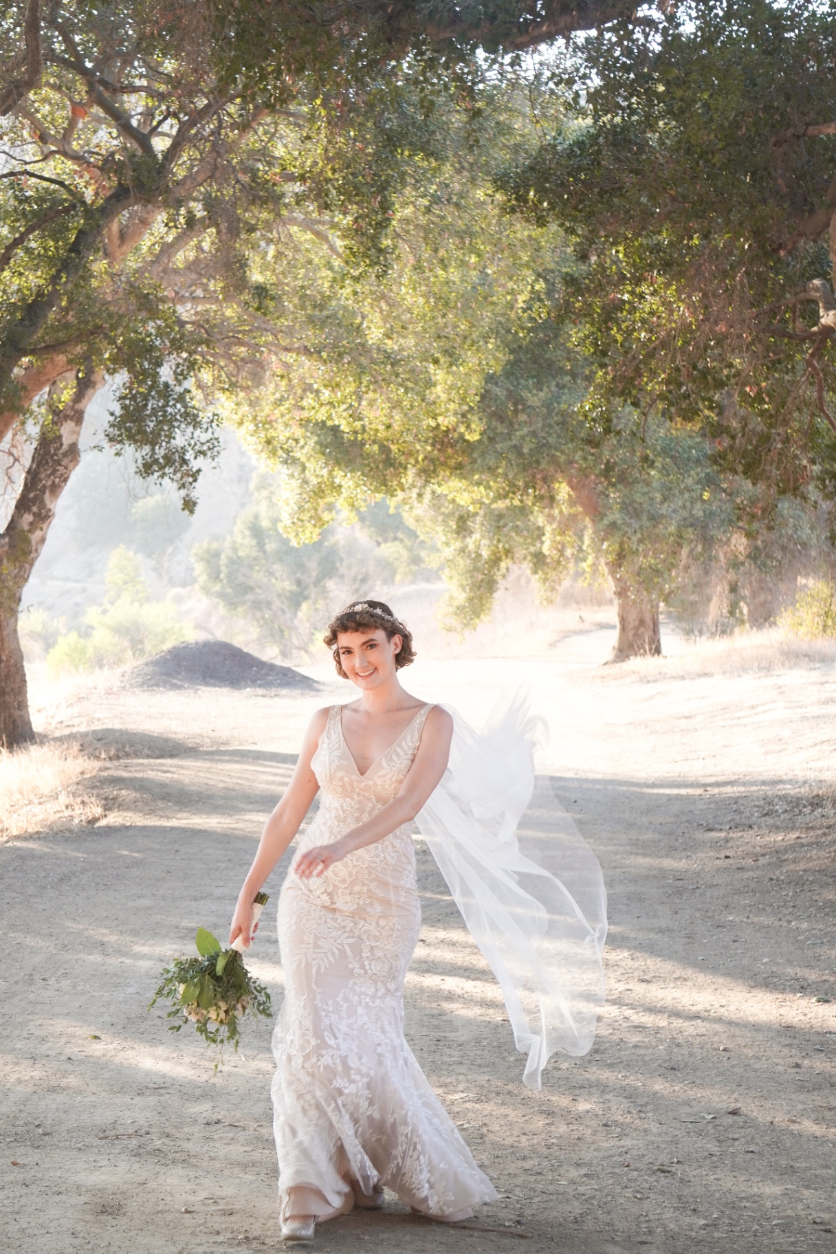 Bride in a lace gown with flowing veil and bouquet under California oak trees on a Los Angeles wedding day
