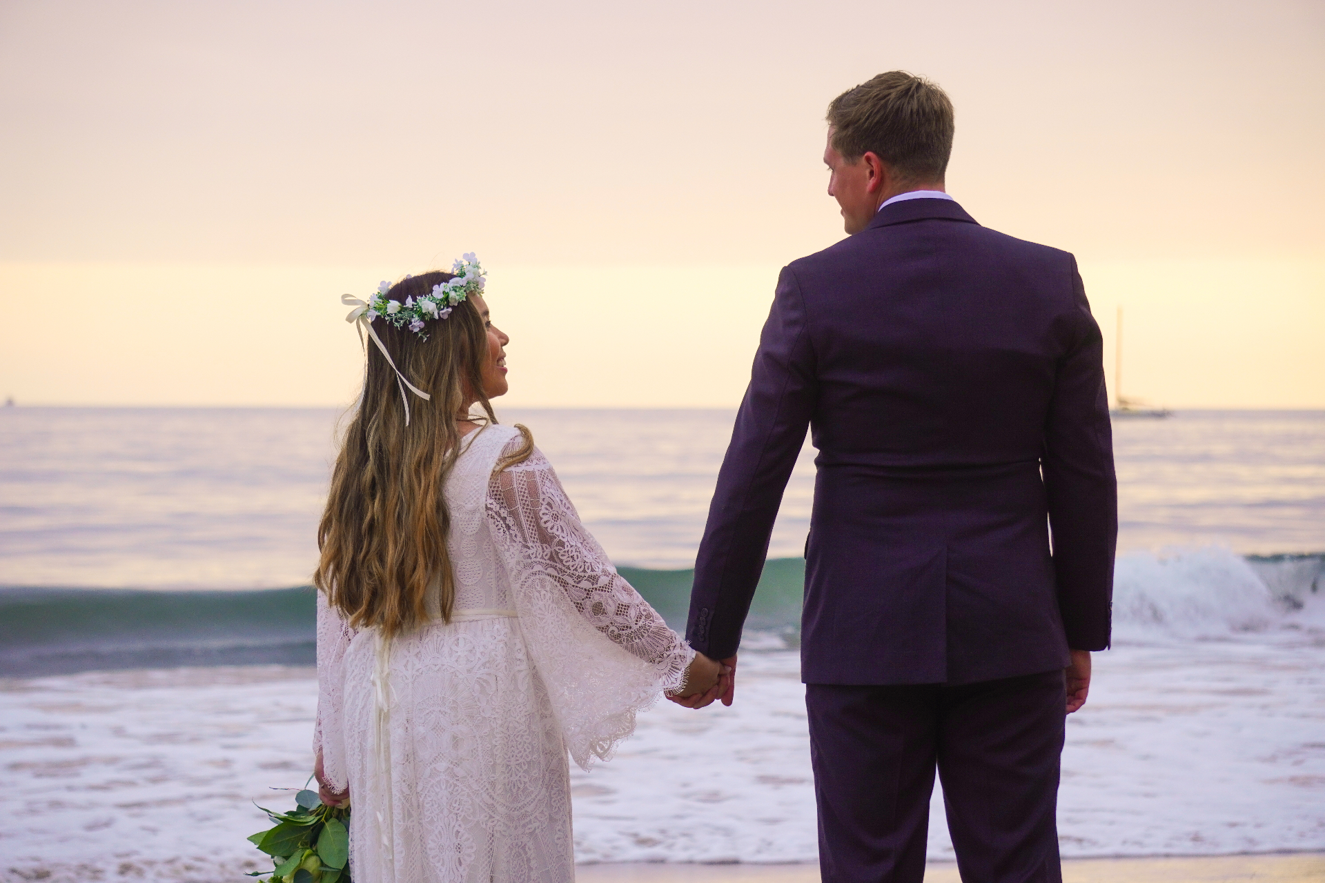 Bride in lace gown with floral crown and groom holding hands at sunset on a Hawaii beach