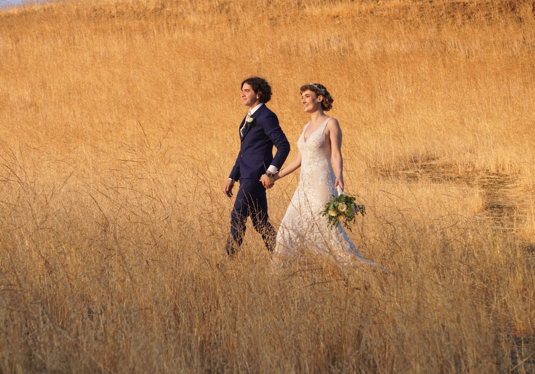 Malibu wedding couple walking through golden grass hills holding a white rose bouquet at sunset